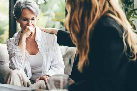 In einem modernen Wohnzimmer unterstützt eine junge Frau eine ältere, sichtlich nachdenkliche Frau mit einer mitfühlenden Geste während eines Gesprächs.