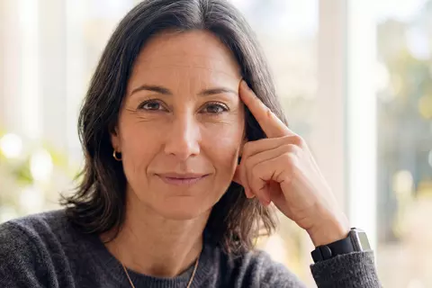 Porträt einer Frau mittleren Alters, die den Betrachter direkt anblickt und ihren Kopf auf ihrer Hand mit gestrecktem Zeigefinger abstützt.