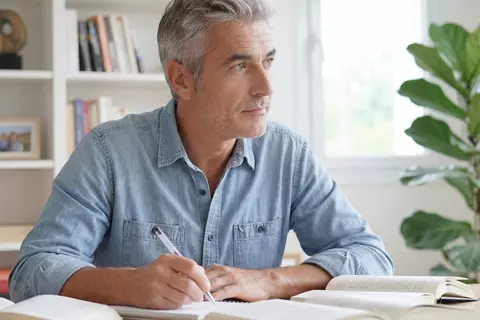 Nachdenklicher Mann studiert Unterlagen am Schreibtisch vor einem Regal voller Bücher in einem hellen Raum.