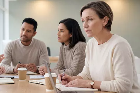 Eine diverse Gruppe von fünf Kollegen sitzt an einem Konferenztisch und hört einem sprechenden Mann aufmerksam zu. Eine Frau hat jedoch Probleme, sich zu konzentrieren.