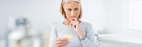 Eine ältere Dame hält in der einen Hand einen Notizzettel und legt die andere Hand nachdenklich an ihr Kinn