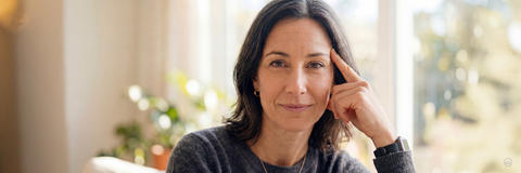 Porträt einer Frau mittleren Alters, die den Betrachter direkt anblickt und ihren Kopf auf ihrer Hand mit gestrecktem Zeigefinger abstützt.