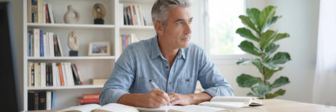 Nachdenklicher Mann studiert Unterlagen am Schreibtisch vor einem Regal voller Bücher in einem hellen Raum.