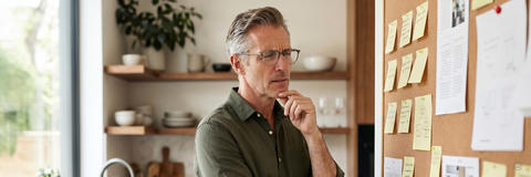 Mann mit Brille betrachtet konzentriert eine Pinnwand voller Notizen in einer modernen Küche. Er wirkt dabei etwas überfordert.