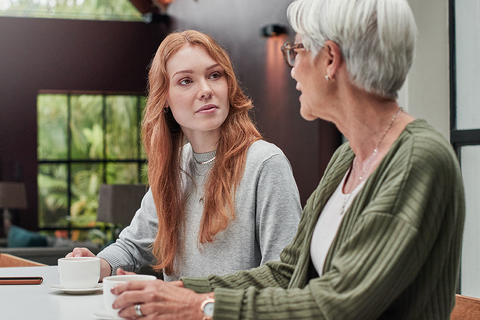 Gespräch zwischen zwei Generationen: Eine Seniorin im grünen Pullover spricht mit einer jungen Frau an einem Tisch.