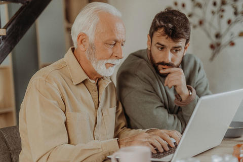 Älterer Mann und jüngerer Mann sitzen zusammen an einem Tisch und schauen konzentriert auf einen Laptop.